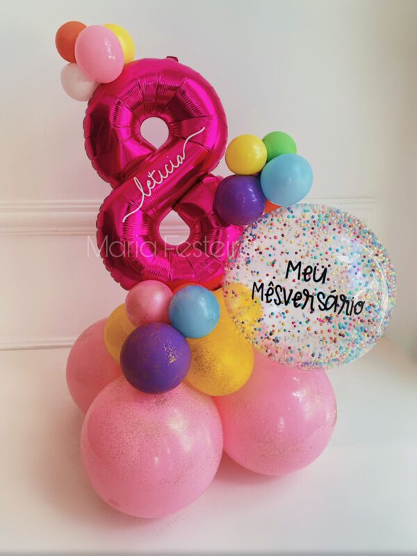 Arranjo de mesversário com balão número metalizado personalizado, balão bolha com confetes coloridos e base decorativa com balões vibrantes.