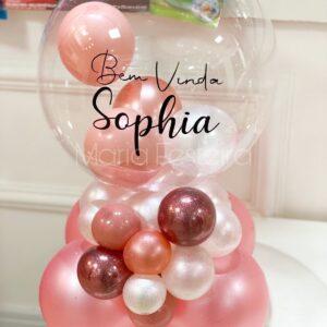 Arranjo de mesa personalizado com bolha transparente decorada com a mensagem 'Bem-vinda Sophia', contendo balões internos em tons de rosa, branco e dourado, ideal para celebrações de boas-vindas ao bebê.