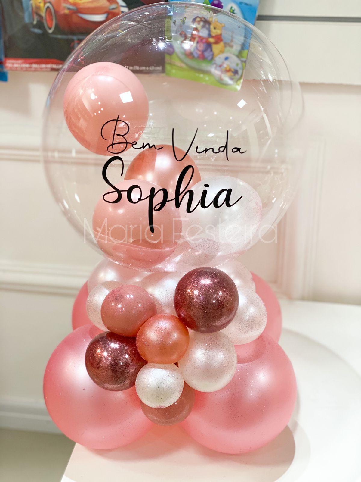 Arranjo de mesa personalizado com bolha transparente decorada com a mensagem 'Bem-vinda Sophia', contendo balões internos em tons de rosa, branco e dourado, ideal para celebrações de boas-vindas ao bebê.