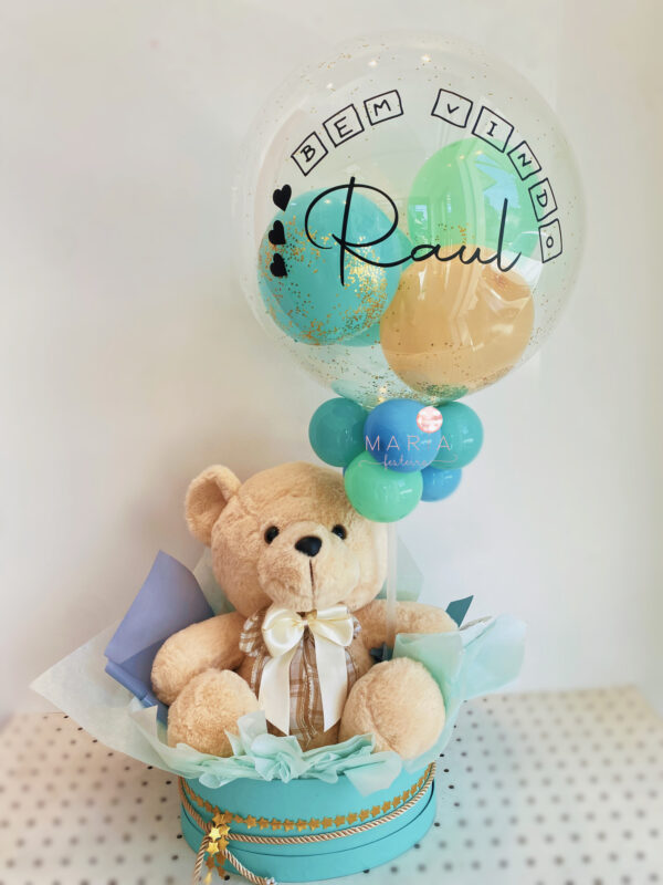 Presente de boas-vindas composto por um ursinho de pelúcia macio e um balão transparente personalizado com nome e mensagem. Balão com confetes e balões internos em cores suaves, fixado em uma base circular decorada com fita de cetim.