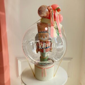 Arranjo natalino com bubble transparente, balão de quebra-nozes dentro, mini balões no topo, laços coloridos e personalização com nome, acomodado em uma elegante base de caixa aberta.