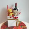 Caixa de Natal com vinho tinto, chocolate Lindt Teddy e bola personalizada transparente recheada com Amanditas, apresentada em caixa branca elegante.